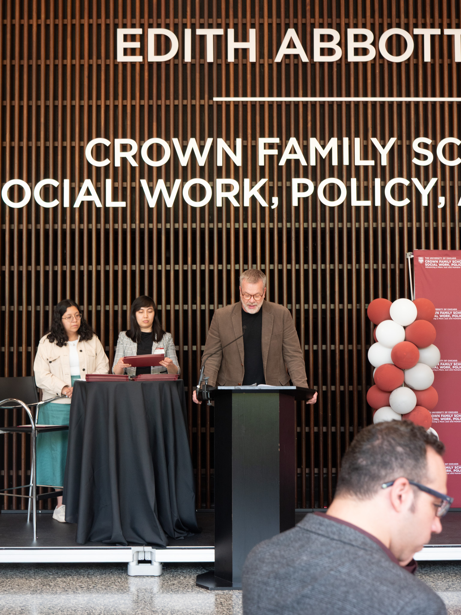Man in brown suit jacket looks down at podium on stage.