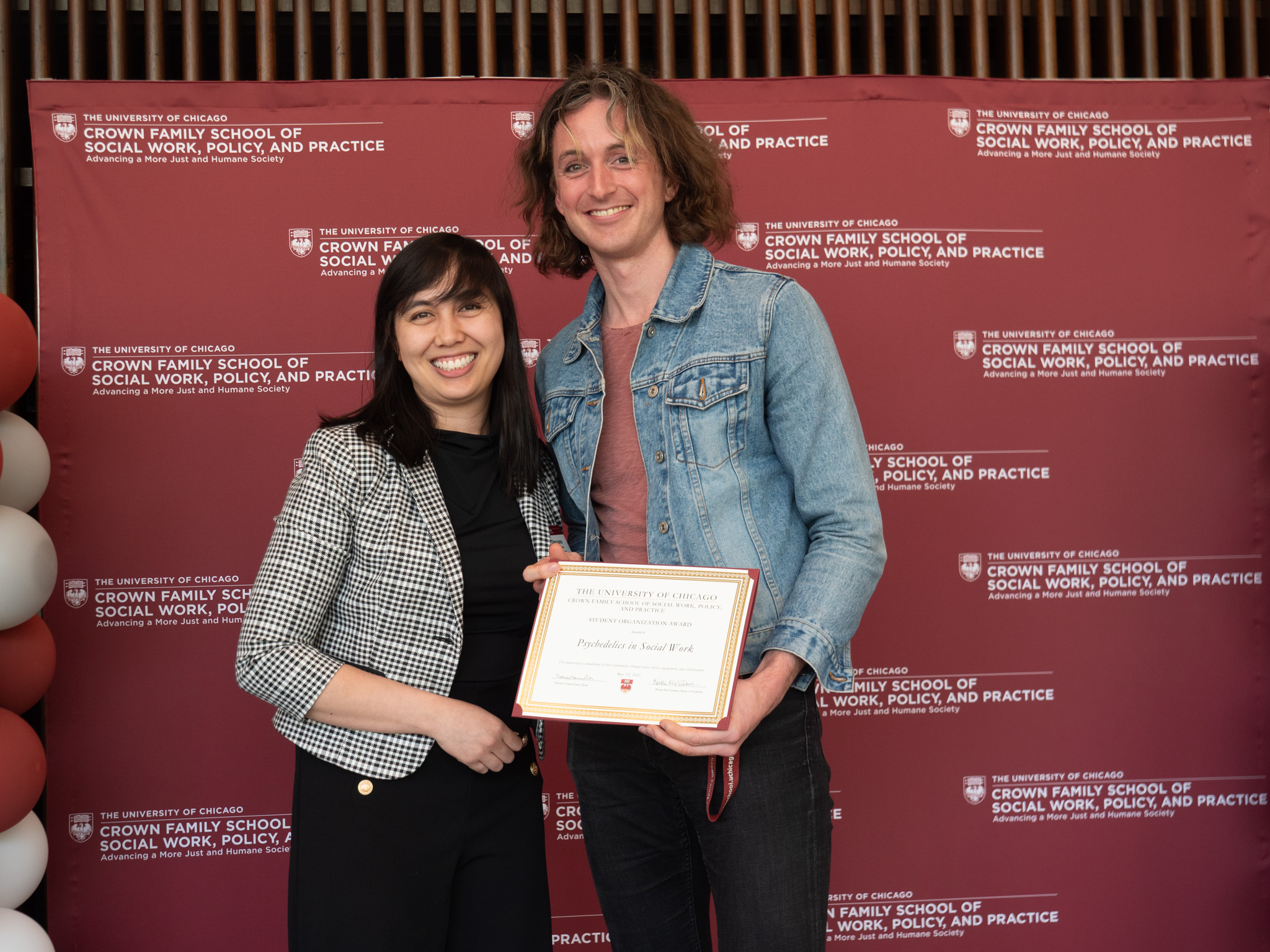 Student awardee wearing blue jean jacket taking picture with woman in black and white blazer  in front of backdrop