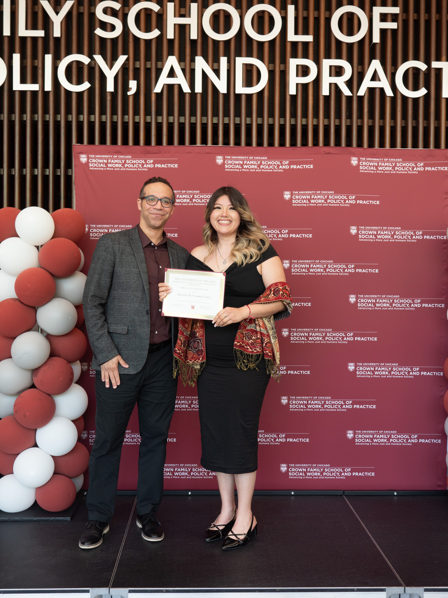 Student awardee wearing black dress and colorful scarf taking picture with man in grey suit jacket in front of backdrop