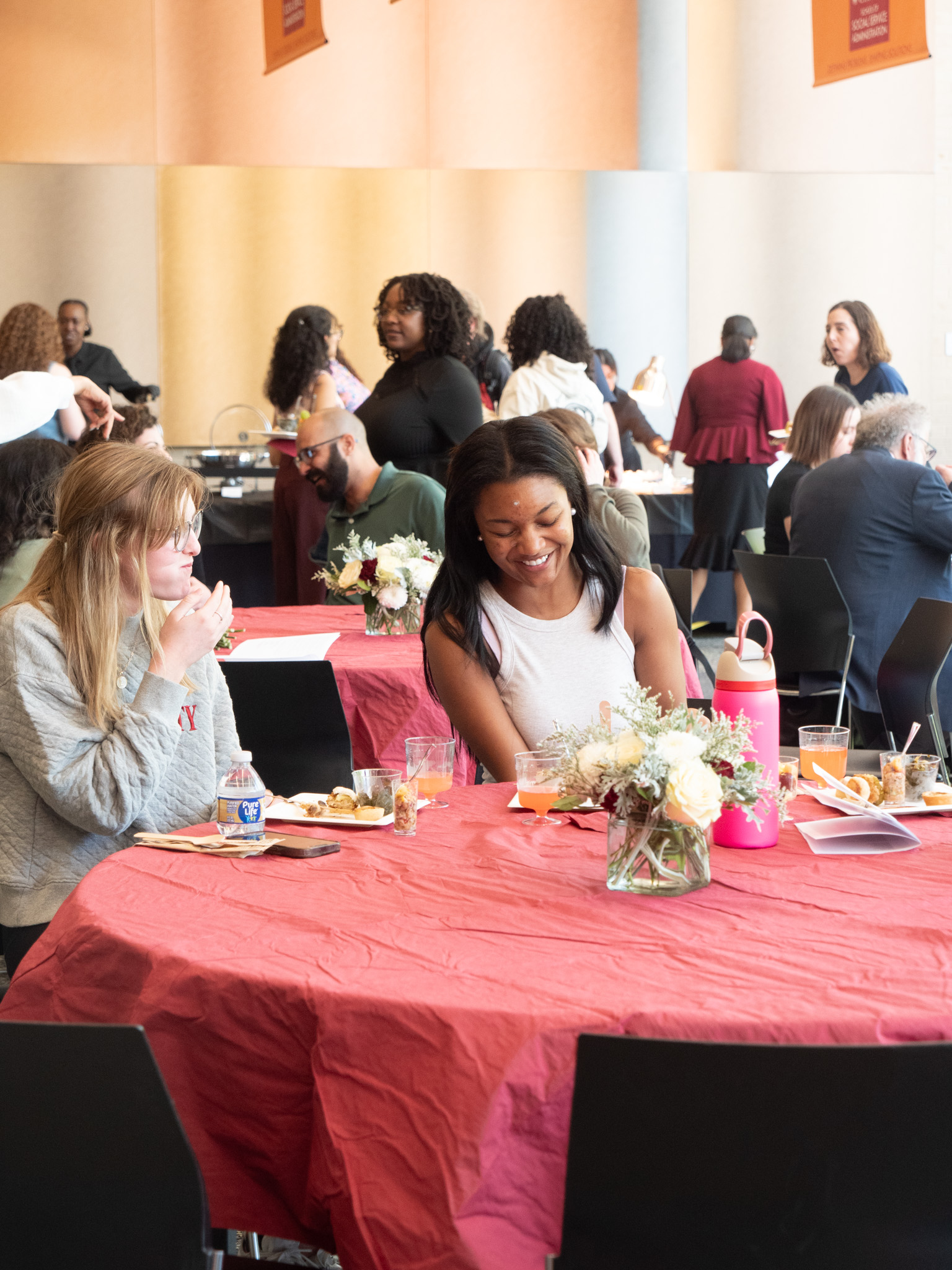 Black woman in white tank top smiling sitting next to white woman in grey sweater eating