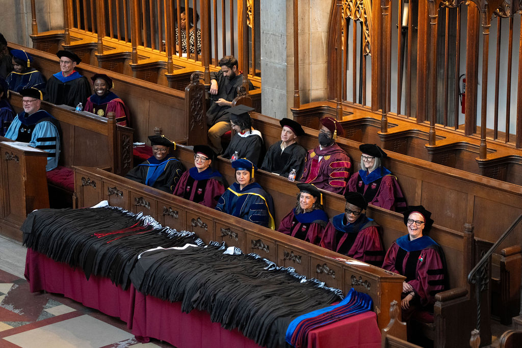 Group of faculty in regalia sitting in pews behind a table of hoods. 