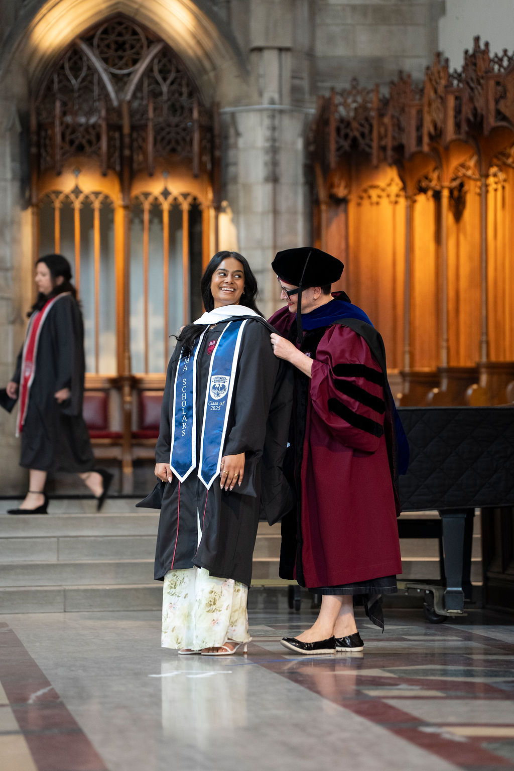 Woman in black robe with blue sash being hooded by a woman in a maroon robe with black hat