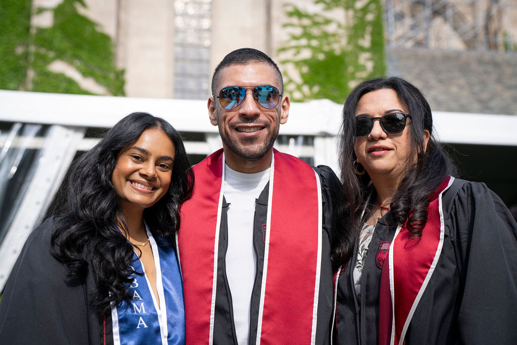 Three graduates in regalia a woman with a blue slash smiling next to a male with sun glasses in the middle with a red and white sash next to a female with sunglasses in regalia 