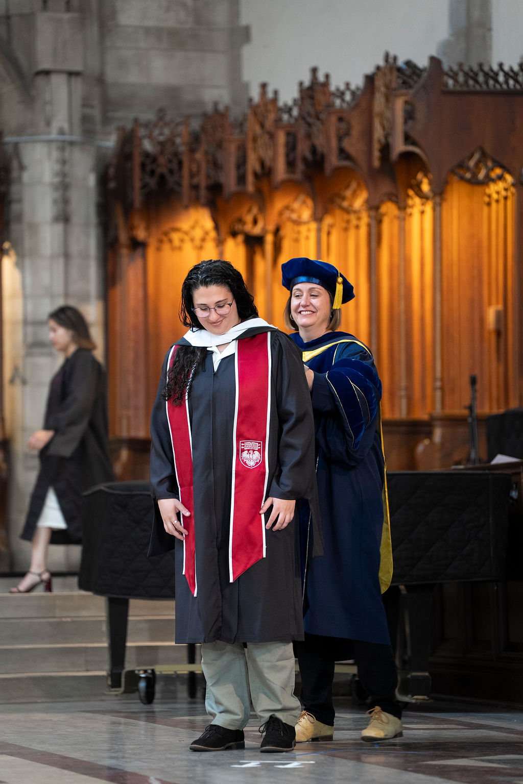 Woman in black regalia with red stole being hooded by a woman in a blue regalia and hat