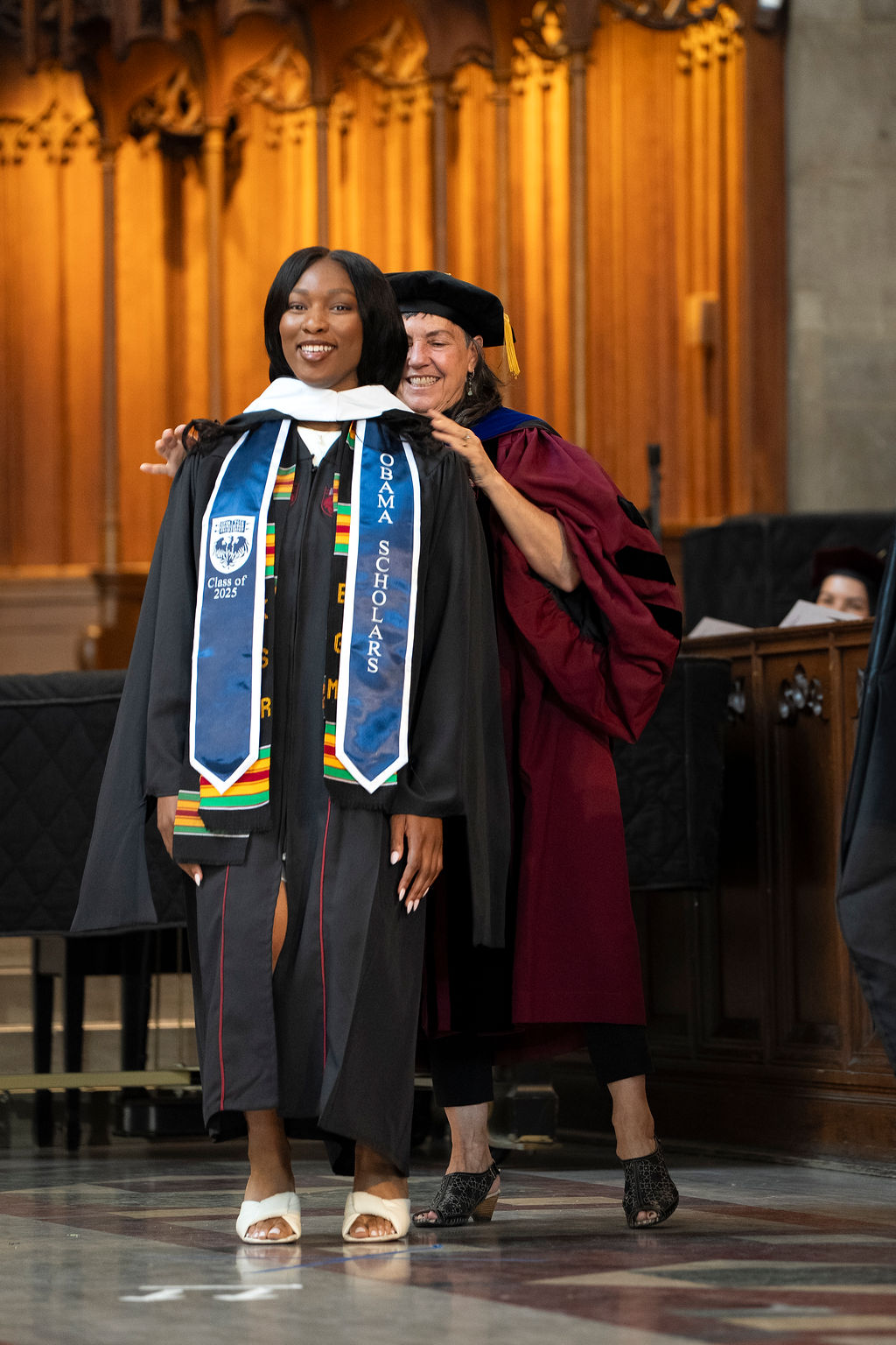 Woman in black regalia with blue stole being hooded by a woman in a maroon robe with black hat