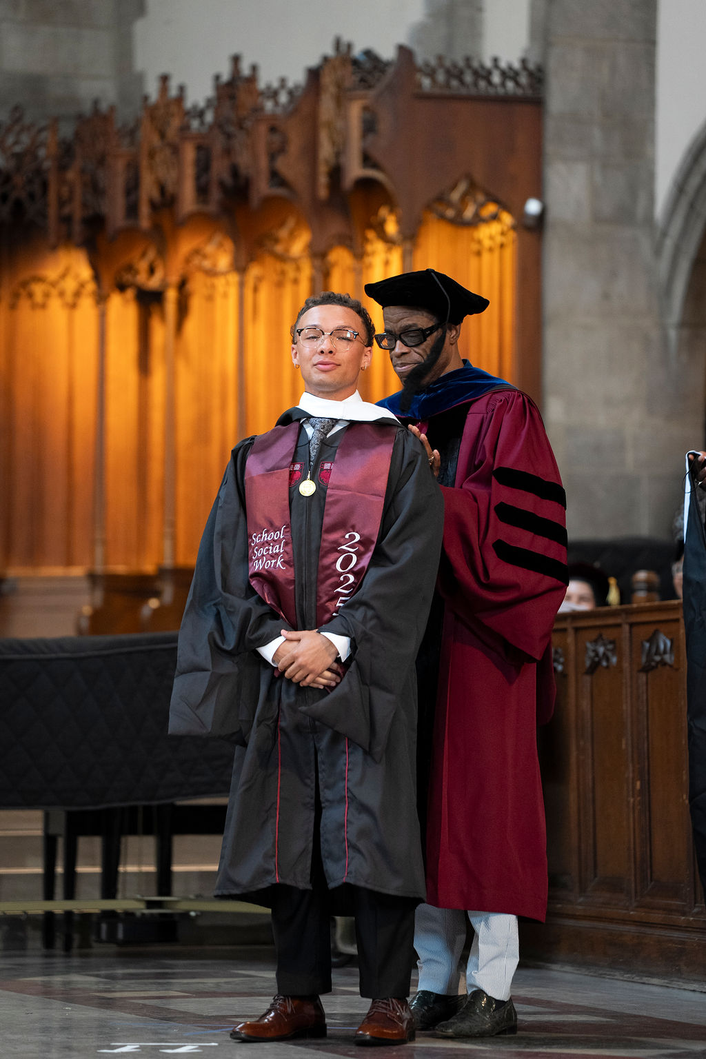 Male in maroon robe with black stripes hooding a male in a black robe with maroon stole