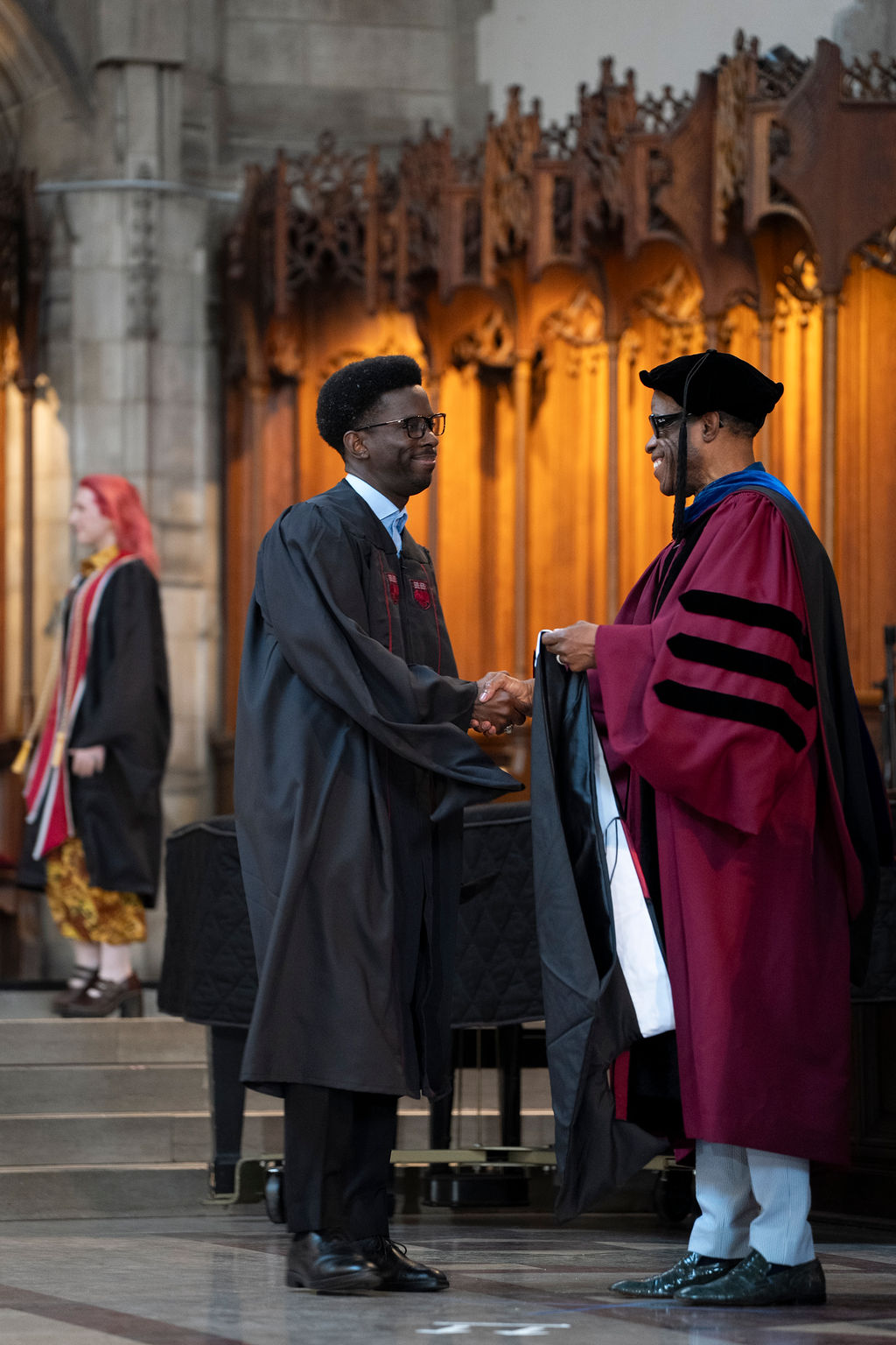 male in black robe shake hands with male in maroon robe with black stripes and hat 