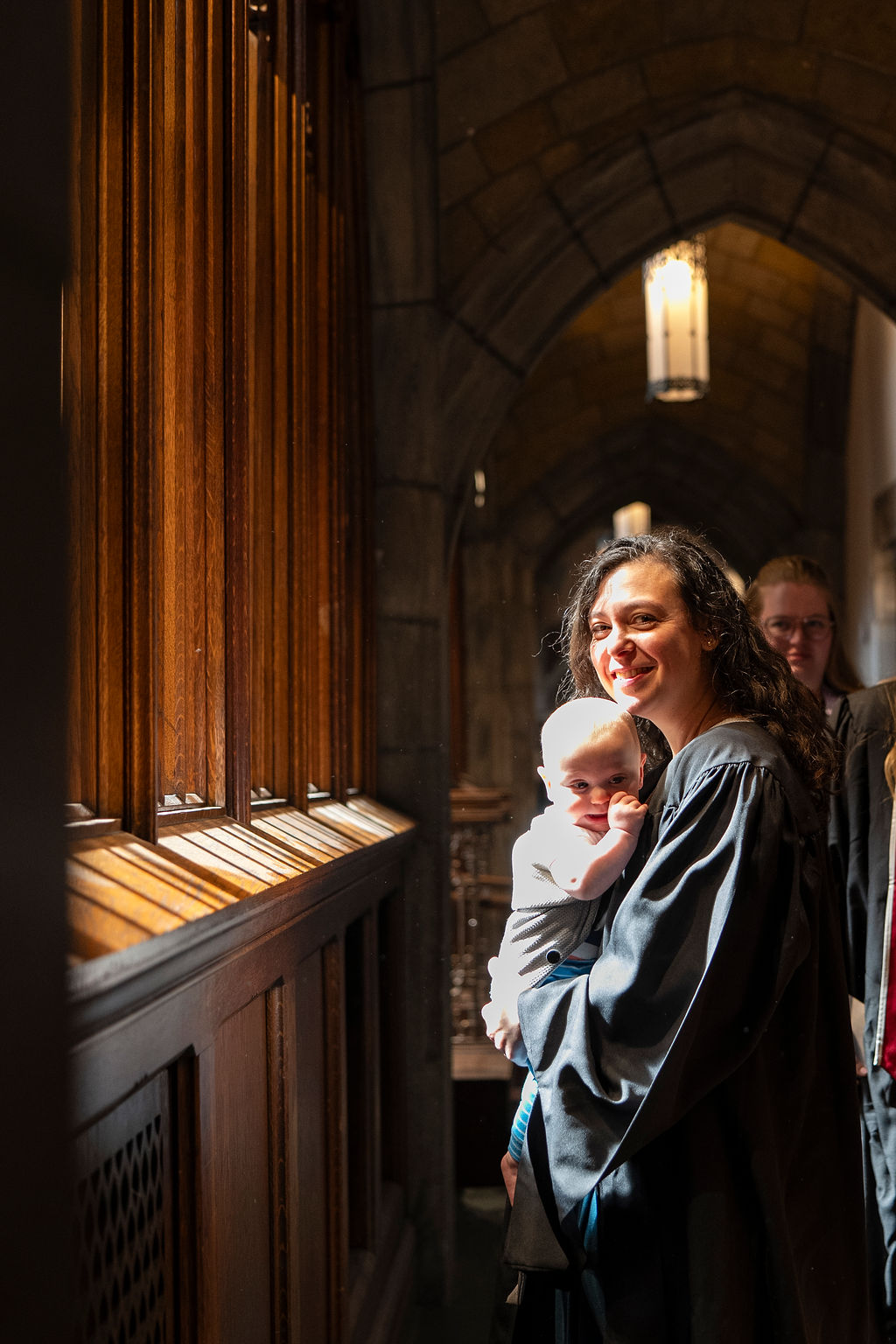 female graduate in regalia smiling while holding a baby with thumb in her mouth 