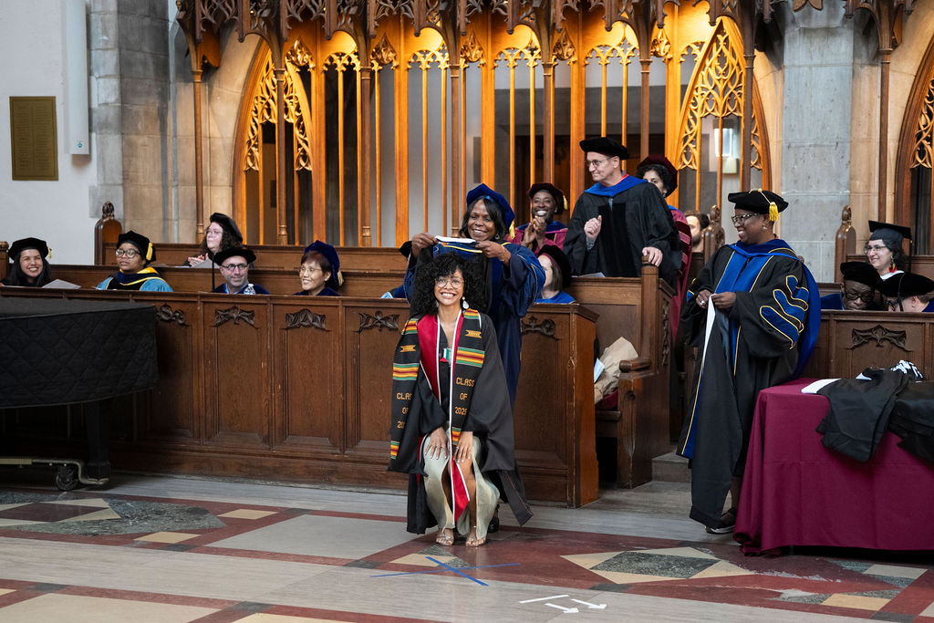 woman in black robe with kinte cloth stole stoops down to be hooded by a woman in a blue regalia while another faculty member in a robe hold the next hood