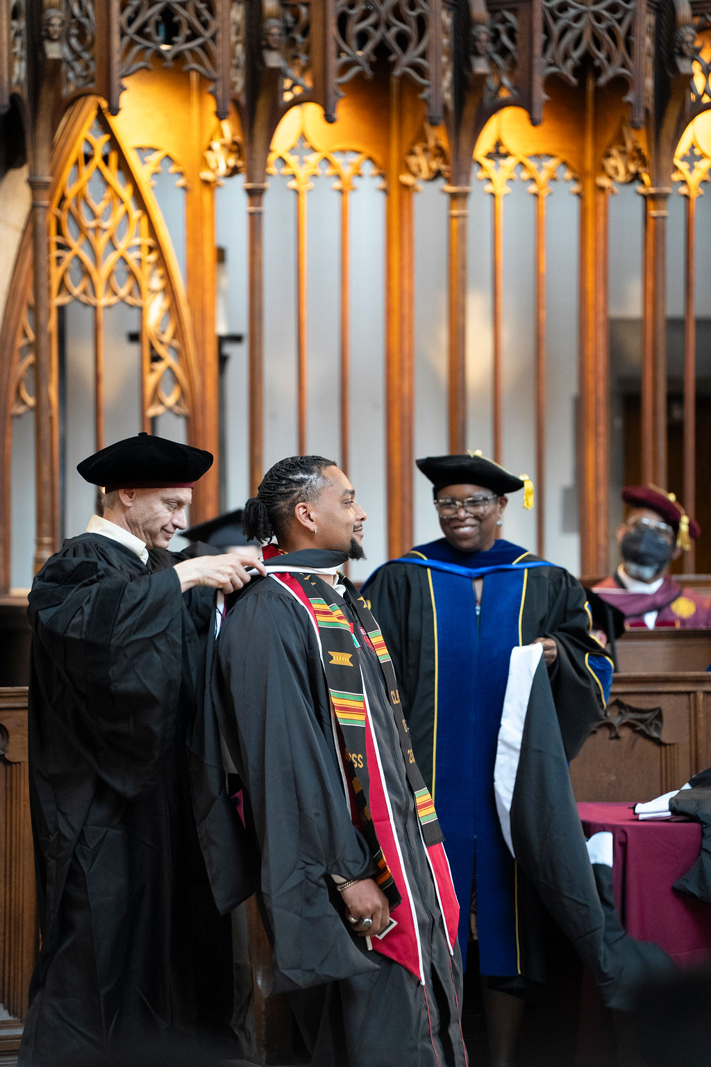 a male graduate in black robe with kinte cloth stole is hooded by a male in black robe and hat while another faculty member in a robe holds the next hood
