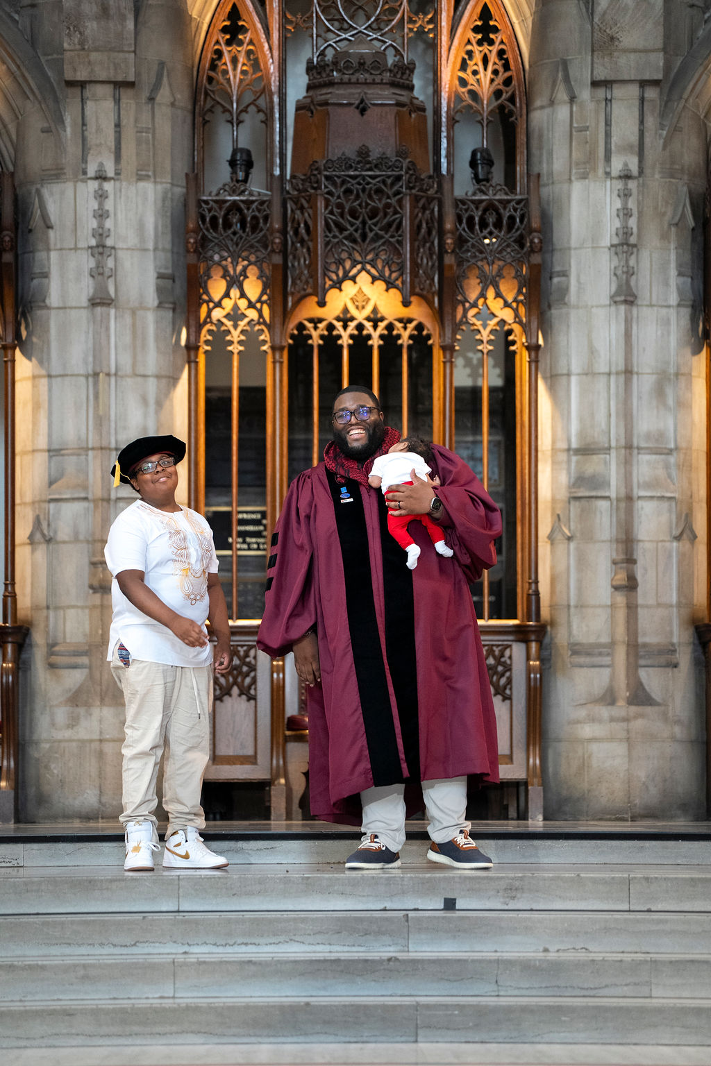 male graduate in maroon regalia holding a newborn baby with kid beaming with pride in khakis and a t-shirt with the graduation tam. 