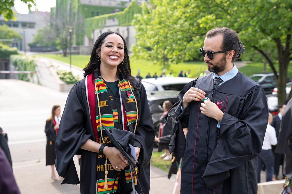 Two graduates in regalia to the left a female smiling with a kente cloth sash with gold cords next to a male with sunglasses in black regalia