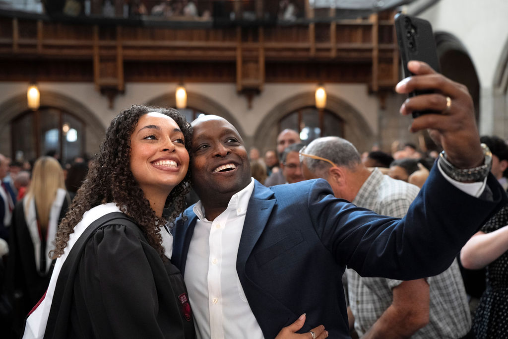 image of a gradate in black regalia with white hood hugging a male while taking a selfie and smiling 