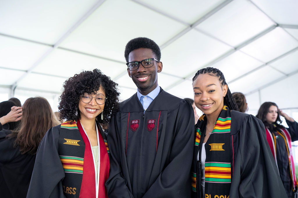 A group of three graduates one female to the left in red with black regalia and African print cloth sash next to a male with black regalia and glasses smiling next to a woman with braids smiling in black regalia with African print  sash