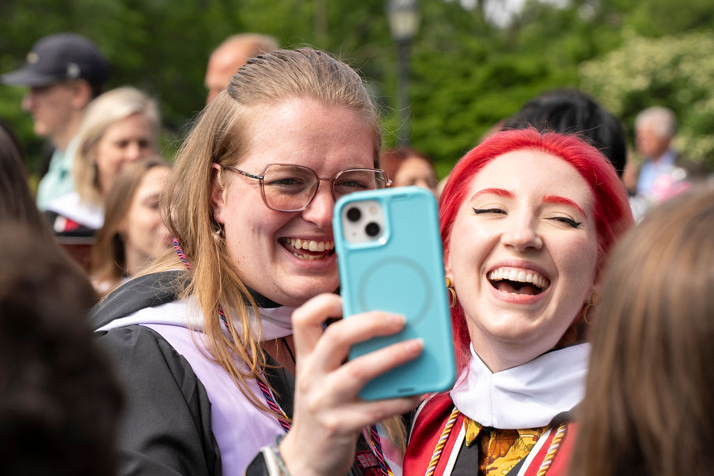 two graduates one with glasses holding a phone with another smiling with red hair take a selfie