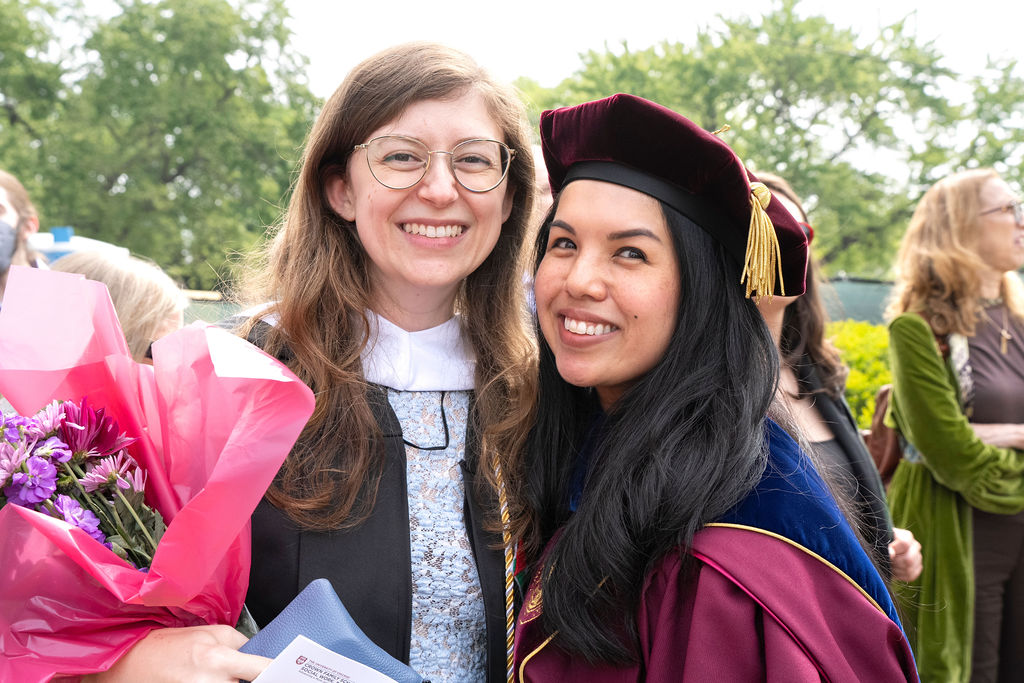 a graduate with glasses in a black robe holding flowers smiles next to a faculty member in maroon regalia smiling