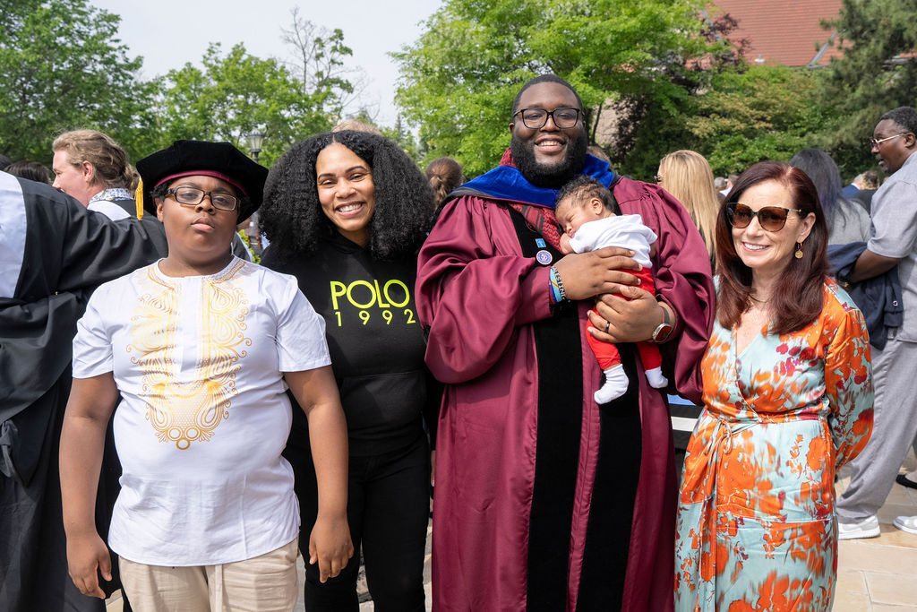 a graduate in a maroon robe holding a newborn stands next to a woman in a flower dress and sunglasses with red hair and another woman with black on with wavy hair who stands next to a kid in the graduation tam with glasses