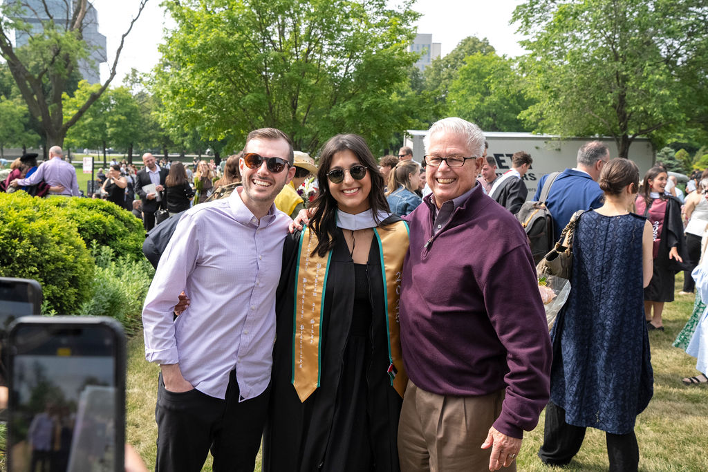 a graduate in a black robe with yellow stole and sunglasses stand next to a male in a maroon quarter zip with glasses and a male with a lavender button up shirt smiling with sunglasses