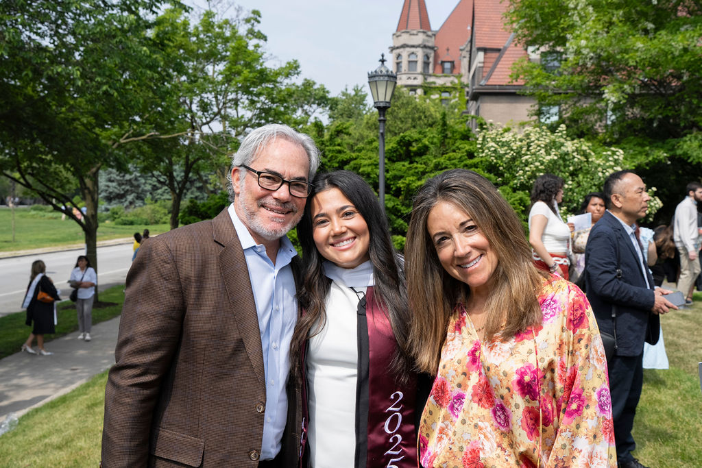 graduate in black robe and maroon stole stands next to a woman in a flowery dress and male in a brown suit jacket with glasses smiing