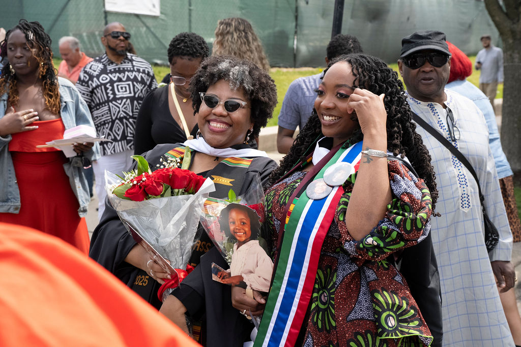 graduate in holding flowers with sunglasses smiling stands next to a woman in a printed dress with green blue and red stole  smiling 