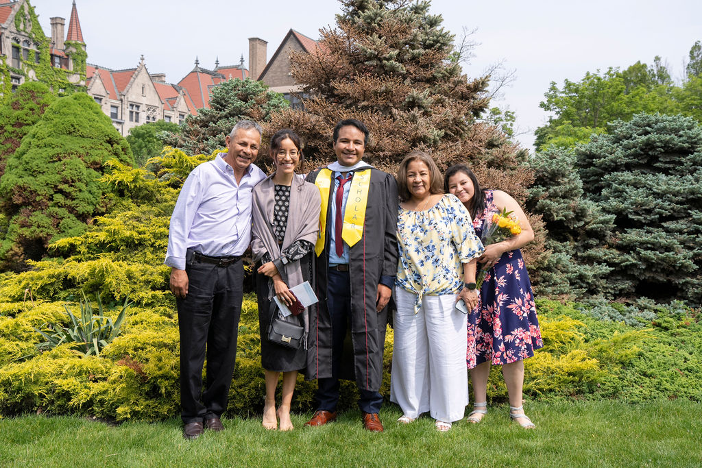 graduate in black robe with yellow stole standing next to four people smiling with joy and pride