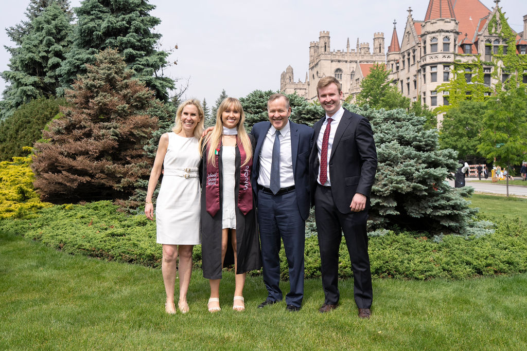 graduate in a black robe with maroon stole standing with three people one woman in white dress to her left and two men in suits to her right with the UChicago buildings in the background