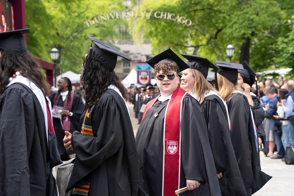 group of graduates in black robes line up with one looking and smiling in a red stole and sunglasses smiling. 