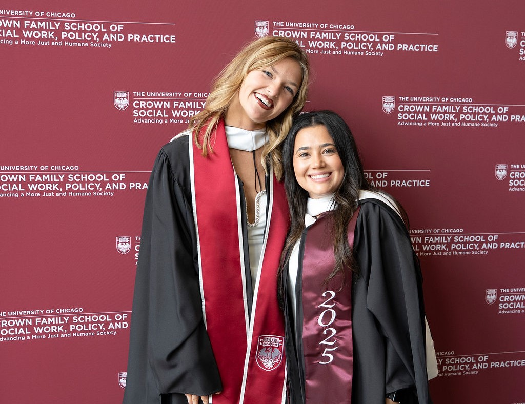 two female graduates in regalia smile in front of a step and repeat that says Crown Family School