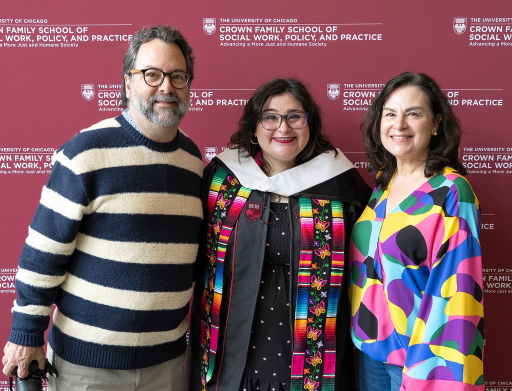 male in striped sweater standing next to a graduate in black regalia with colorful stole next to a woman in a rainbow colored blouse smiling. 