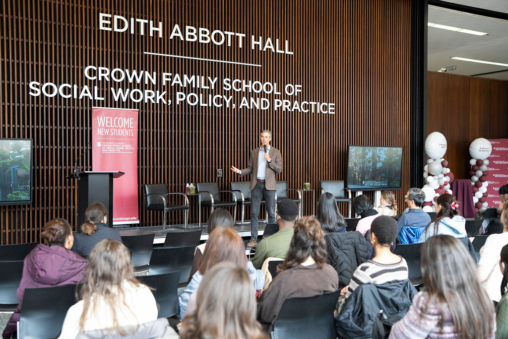 Male in brown suit jacket, blue shirt and black pants holding a microphone speaking to a large group