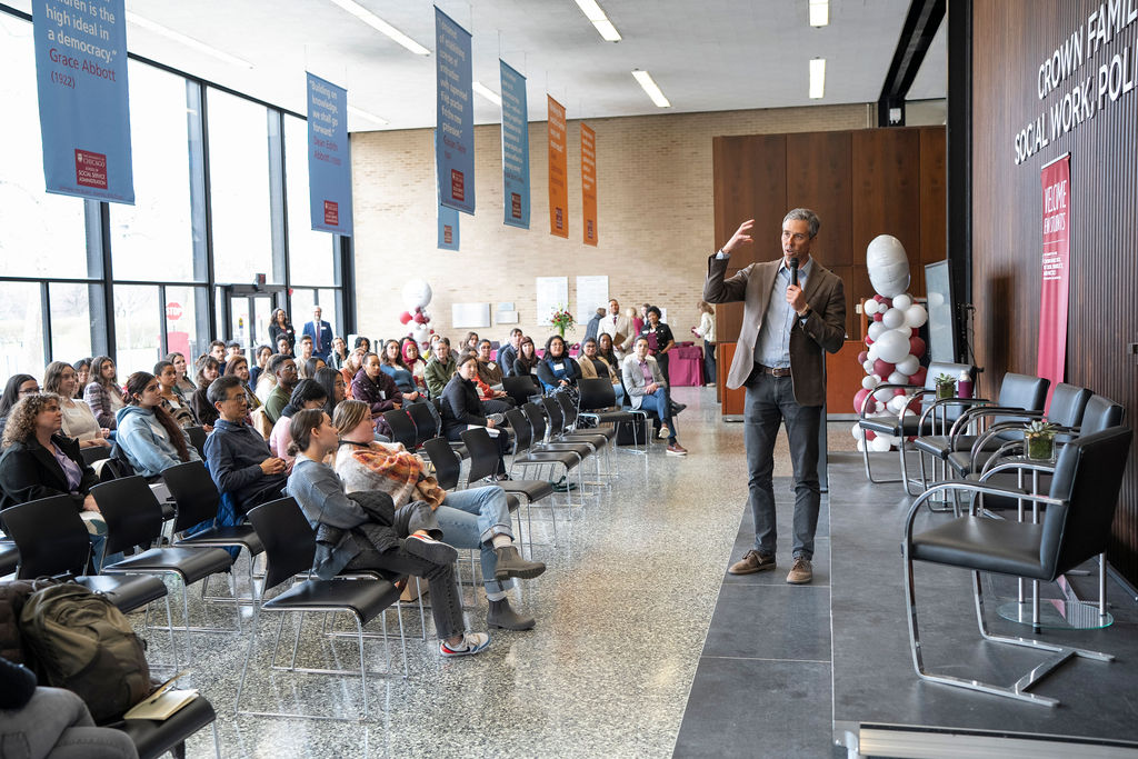 Male in brown suit jacket, blue shirt and black pants holding a microphone speaking to a large group