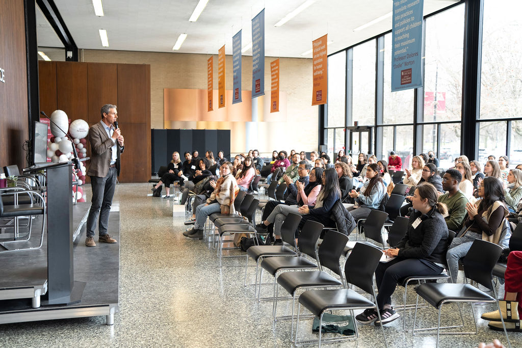 Male in brown suit jacket, blue shirt and black pants holding a microphone speaking to a large group