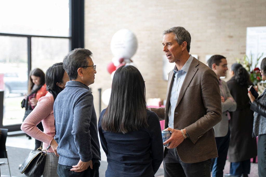 Male in brown suit jacket, blue shirt and black pants speaking to a group of three people