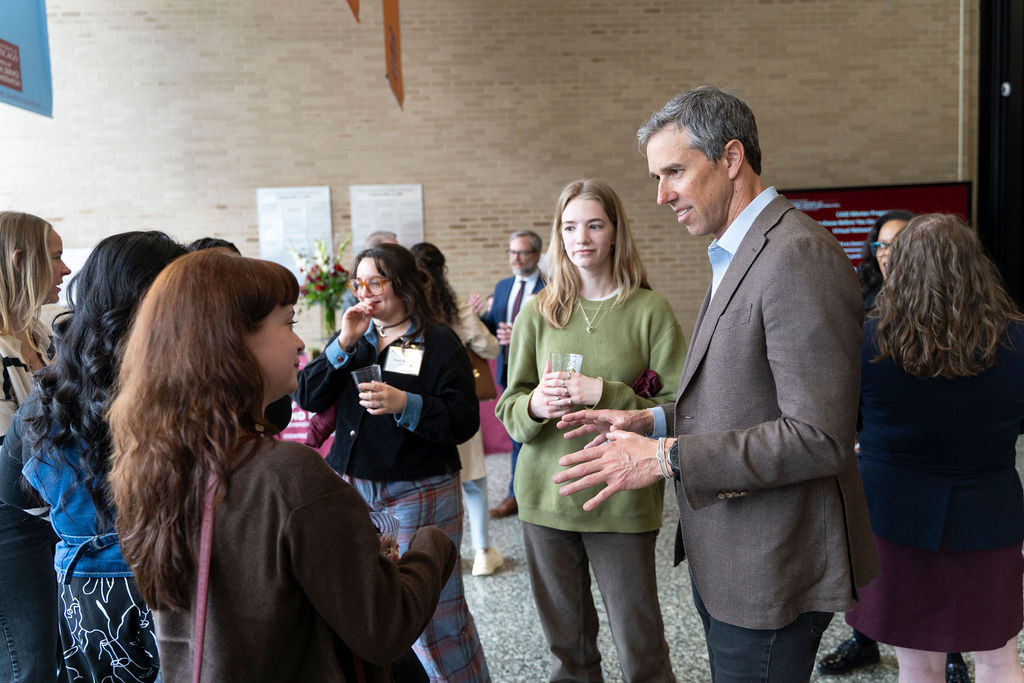 Male in a brown suit jacket standing speaking with a small group of people in a semi circle