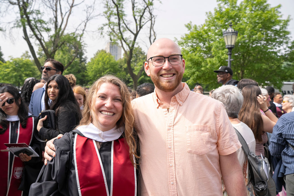 Image of a female graduate in black regalia and red stole standing and smiling next to a male in a salmon colored button up shirt with glasses and red haired beard and mustache