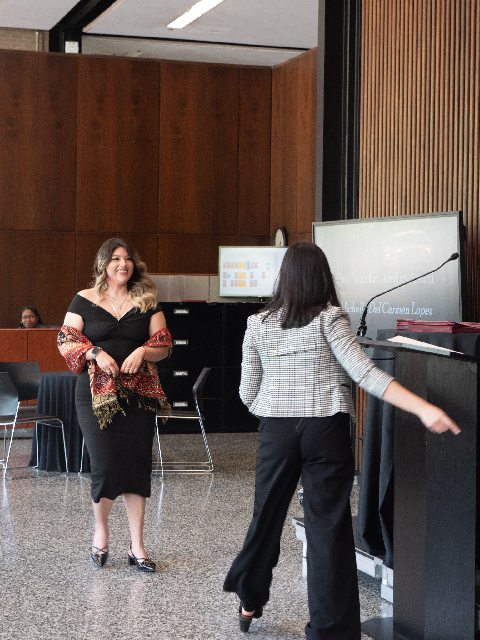 Woman in black dress walking to the podium with another woman in black pants and checked jacket