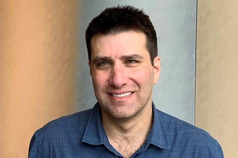 Aaron Gottlieb, a male-presenting person, smiles towards the camera against a grey and orange background.