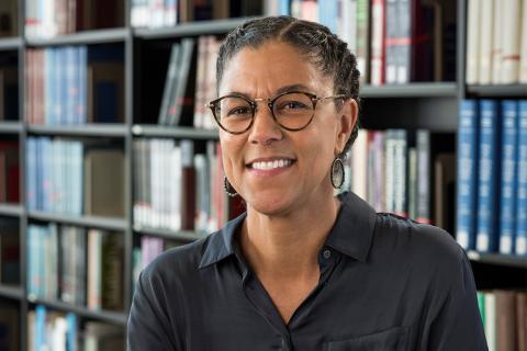 Gina E. Miranda Samuels, a female-presenting person, smiles towards the camera in a library.