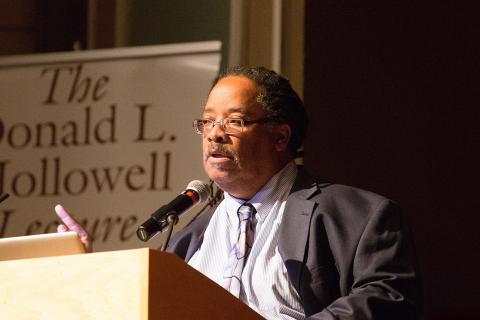 Llewellyn Cornelius speaking at a podium. He has glasses and is wearing a gray suit.