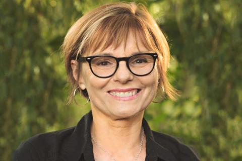Karen Graham headshot; Karen has short light brown hair, wears black framed glasses and a black collared shirt and poses in front of a green leafy background