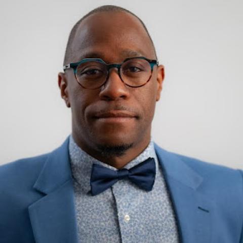 Photo shows a headshot of a man with glasses, wearing a jackets, shirt and bowtie in different shades of blue