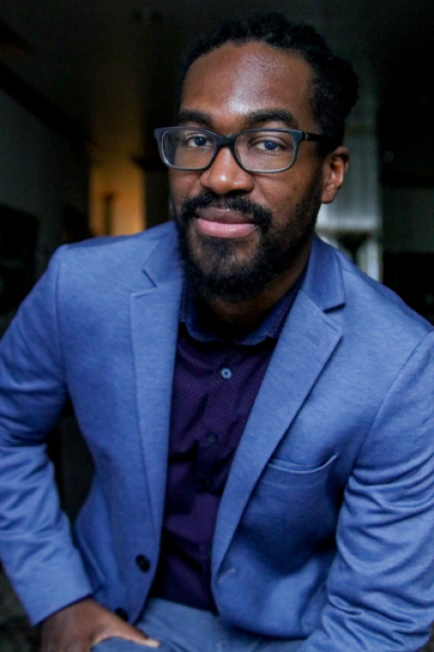 Photo shows a headshot of a man with black glasses, black shirt and a blue blazer