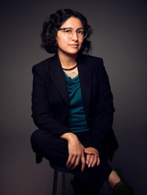 woman in black jacket with teal blouse with glasses and dark hair sitting with crossed leg