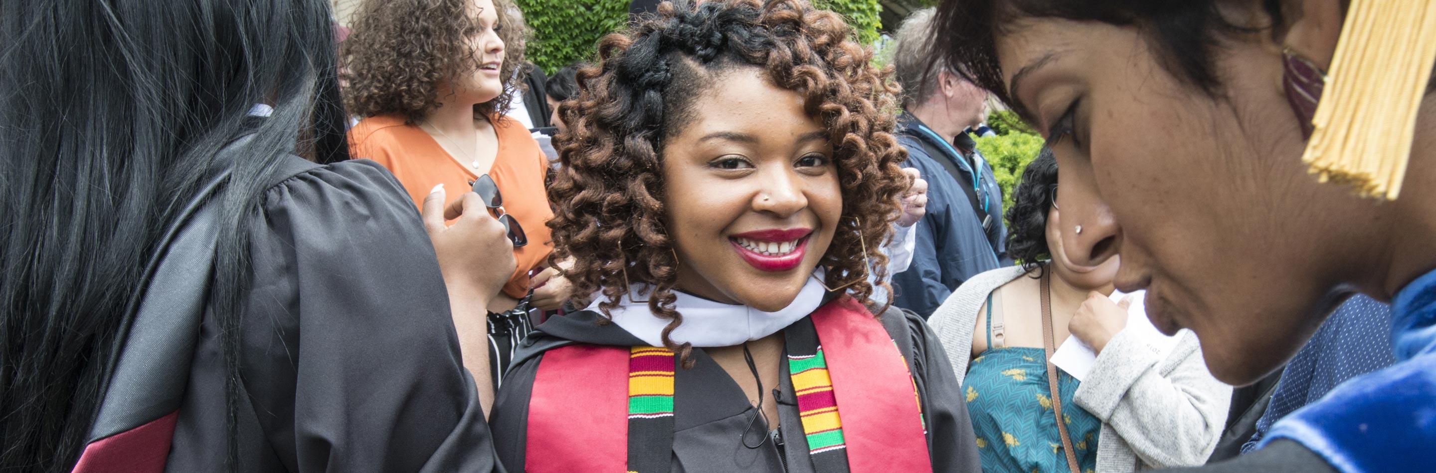 A dark-skinned, curly-haired person in graduation regalia smiling in a group of graduates outside