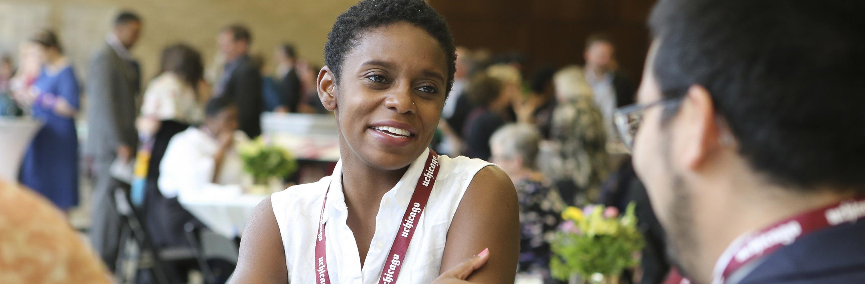 A person smiling, wearing a U Chicago lanyard, talking to somebody at a meal table