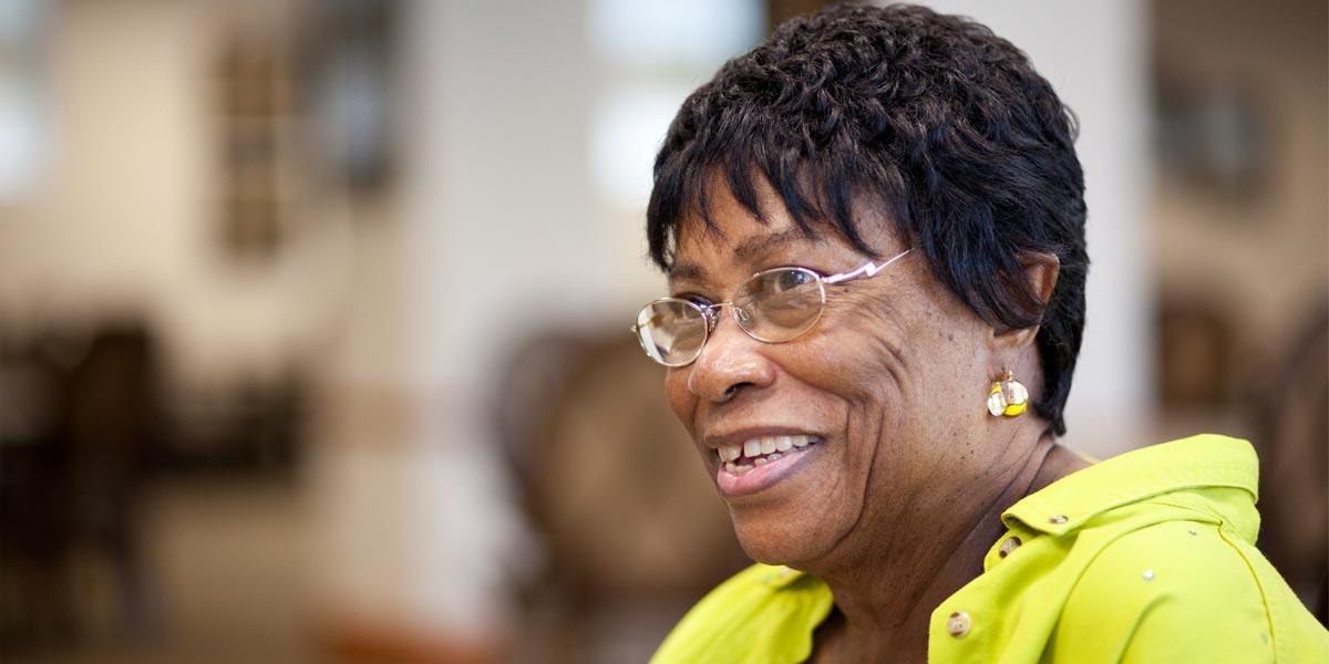 Older African American woman with glasses, gold earrings, and yellow shirt smiles brightly at the camera 