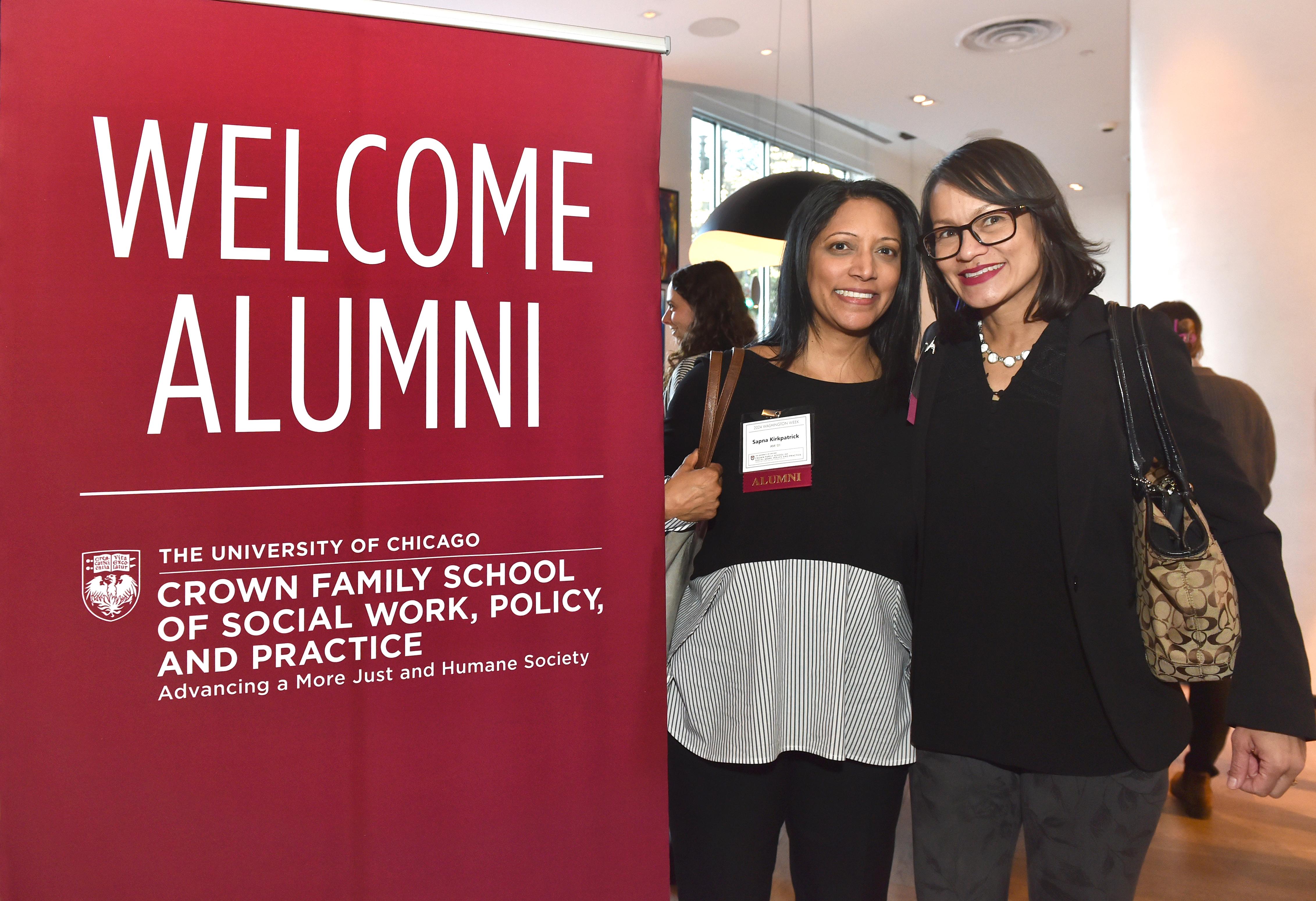 Image of two women in black smiling next to a welcome alum sign