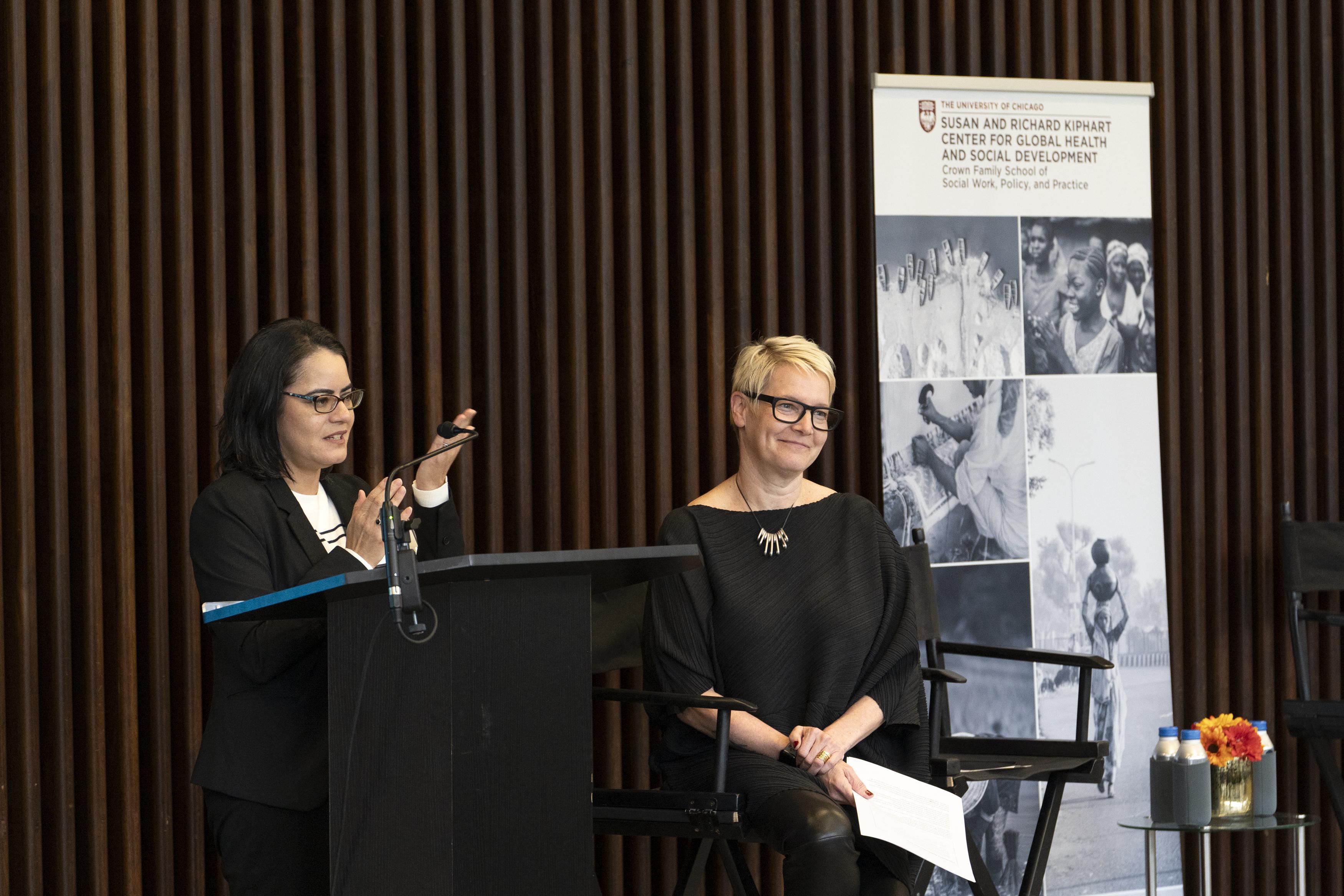 One woman in black standing at podium talking with another woman sitting down smiling