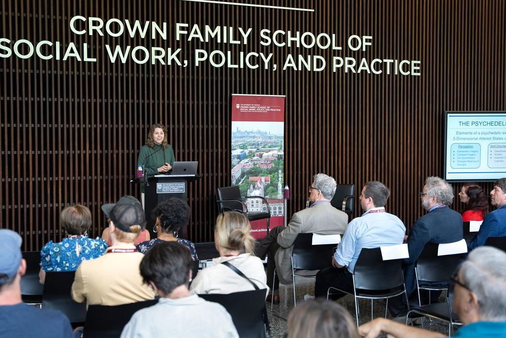 Woman in green shirt and jacket stands at podium speaking to an audience