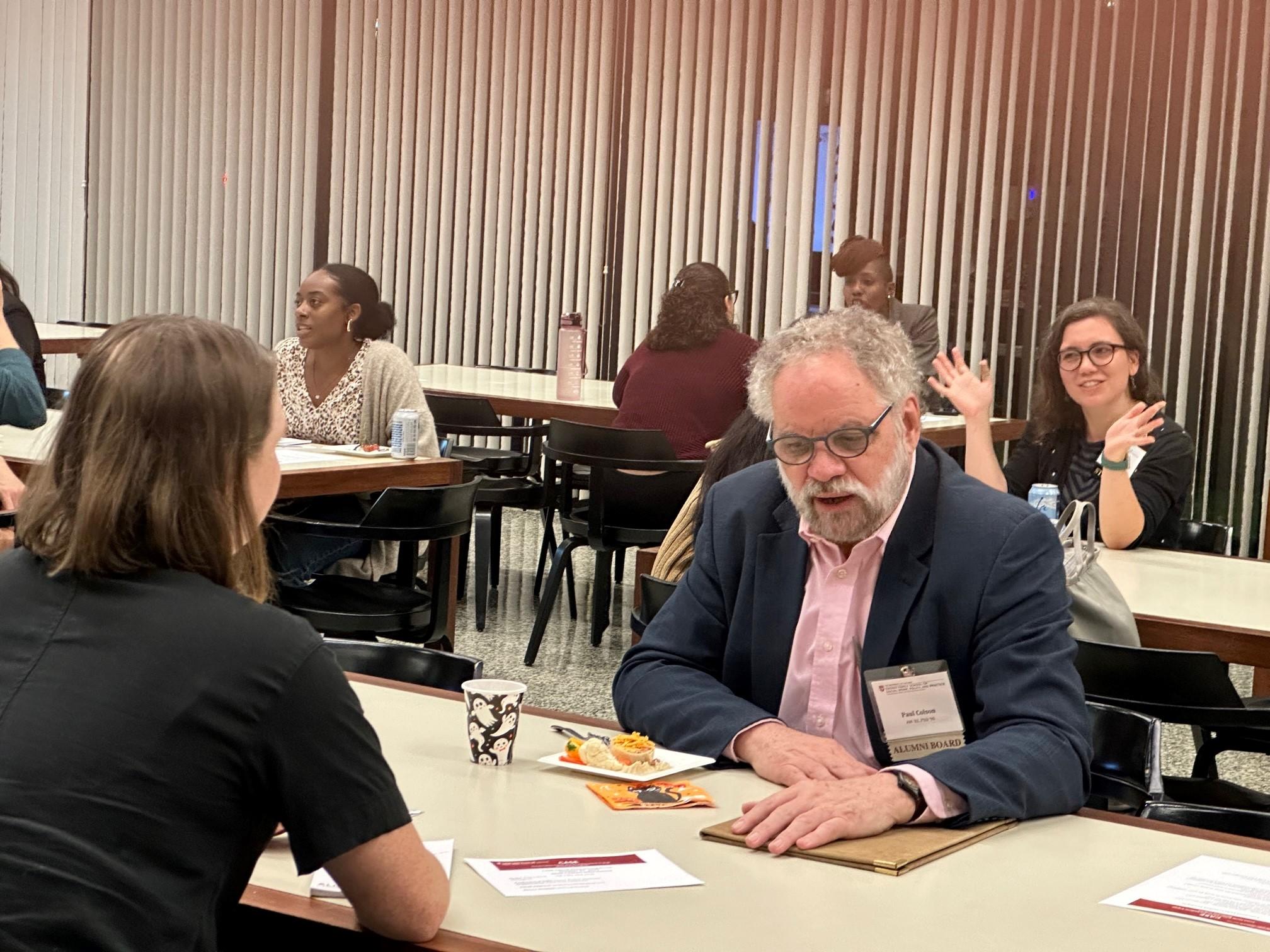 The photo shows a room of multiple tables each as two people sitting across from one another engaged in one on one conversations. 